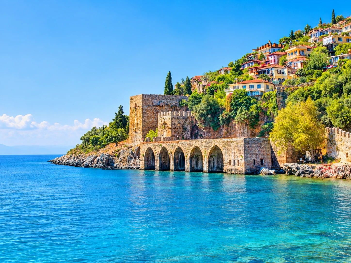 Det seljukiske verftet Tersane med fem steinbuer ved vannkanten i Alanya, med borgmur, tårn og fargerike hus i den grønne skråningen over turkisblått hav